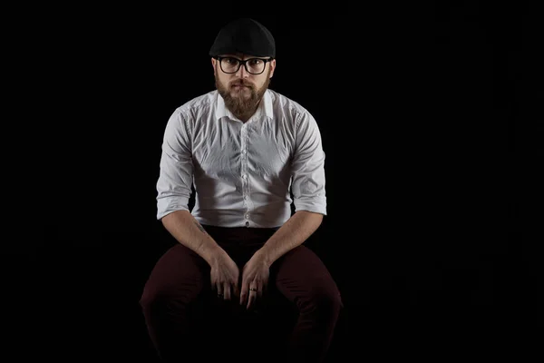 Portrait of a young man in glasses with a beard and a mustache in a black cap and a white shirt, A dark studio background