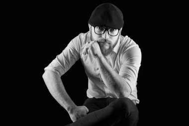 genç adam gözlüklü sakallı ve bıyıklı bir siyah kap ve dizini, bir karanlık studio arka plan karşı yaslanmış onun sandalyede oturan bir beyaz gömlek çok sakallı