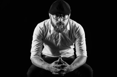 Genç oldukça büyük sakallı adam gözlüklü sakallı ve bıyıklı bir siyah şapka ve beyaz bir gömlek bir karanlık studio arka plan doğrudan ciddi ve bilerek, bir sandalye üzerinde oturur
