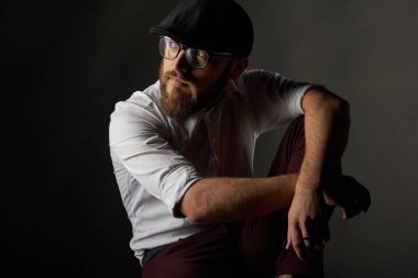 genç adam gözlüklü sakallı ve bıyıklı bir siyah kap ve dizini, bir karanlık studio arka plan karşı yaslanmış onun sandalyede oturan bir beyaz gömlek çok sakallı