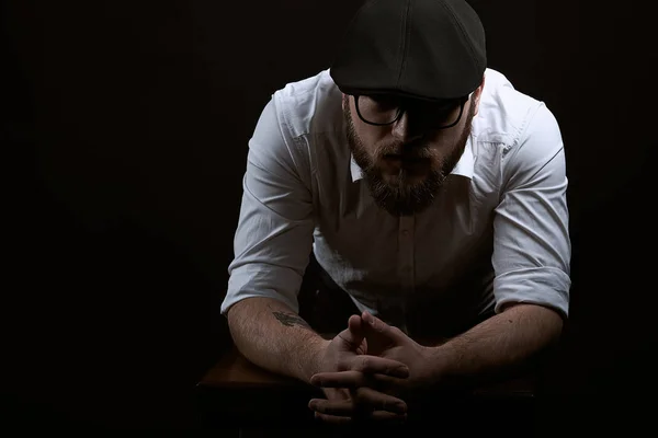 young handsome man in the glasses of struggle and mustache in a black cap and a white shirt conceived bent on his hands, a dark studio background