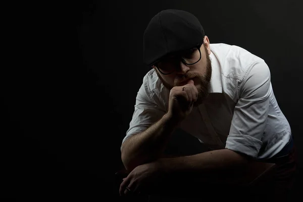 young handsome man in the glasses of struggle and mustache in a black cap and a white shirt conceived bent on his hands, a dark studio background