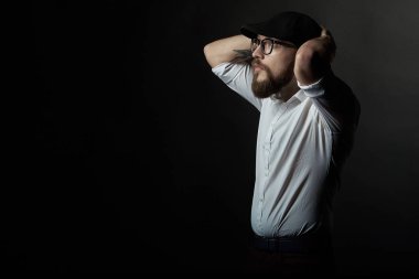 genç yakışıklı adam gözlüklü bir sakal ve bıyık bir siyah şapka ve beyaz bir gömlek ile kollarını kafasının arkasında tutar ve yana doğru bir karanlık studio arka plan standları