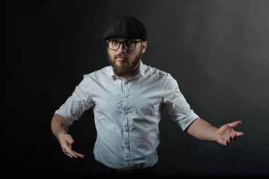 genç yakışıklı adam gözlüklü sakallı ve bıyıklı bir siyah şapka ve beyaz bir gömlek onun ellerini onun karanlık studio arka plan önünde tutar 