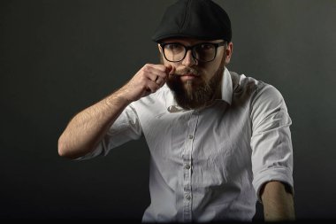 Sakal ve bıyık bir siyah şapka ve beyaz bir gömlek ile gözlüklü genç yakışıklı adam bıyığının eliyle karanlık studio arka plan giderir. 
