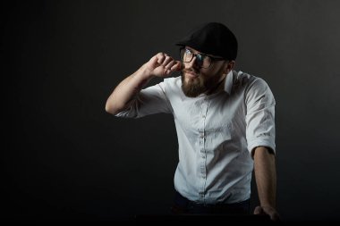 Sakal ve bıyık bir siyah şapka ve beyaz bir gömlek ile gözlüklü genç yakışıklı adam bıyığının eliyle karanlık studio arka plan giderir. 