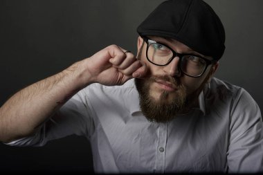 Sakal ve bıyık bir siyah şapka ve beyaz bir gömlek ile gözlüklü genç yakışıklı adam bıyığının eliyle karanlık studio arka plan giderir. 