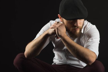 Genç oldukça büyük sakallı adam gözlüklü sakallı ve bıyıklı bir siyah şapka ve beyaz bir gömlek bir karanlık studio arka plan doğrudan ciddi ve bilerek, bir sandalye üzerinde oturur