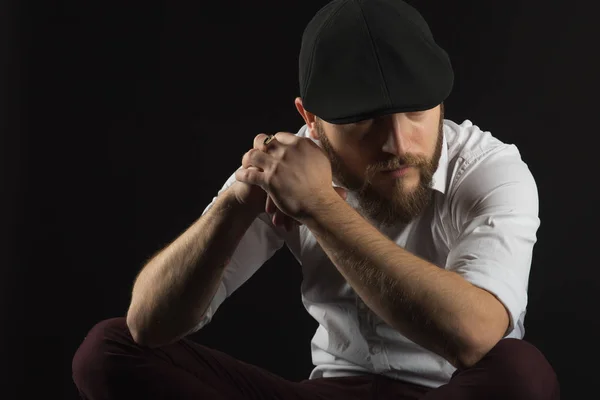 young pretty big bearded man in glasses with a beard and a mustache in a black cap and a white shirt sits on a chair looking directly seriously and purposefully, a dark studio background