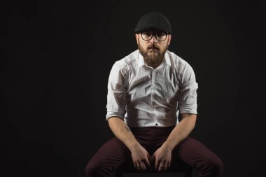 Genç oldukça büyük sakallı adam gözlüklü sakallı ve bıyıklı bir siyah şapka ve beyaz bir gömlek bir karanlık studio arka plan doğrudan ciddi ve bilerek, bir sandalye üzerinde oturur