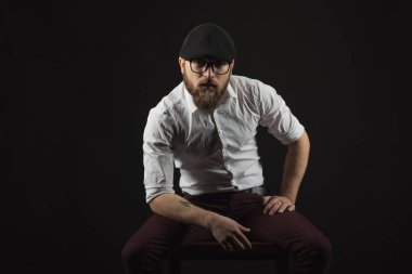 Genç oldukça büyük sakallı adam gözlüklü sakallı ve bıyıklı bir siyah şapka ve beyaz bir gömlek bir karanlık studio arka plan doğrudan ciddi ve bilerek, bir sandalye üzerinde oturur