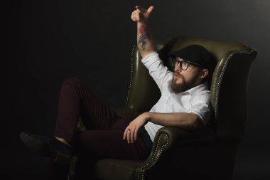 siyah bir kap içinde sakal ve bıyık giyen genç yakışıklı bir adam ve beyaz bir gömlek bir deri yeşil koltuk hamile oturan ve karanlık bir stüdyoda elleriyle jestleri gösteren