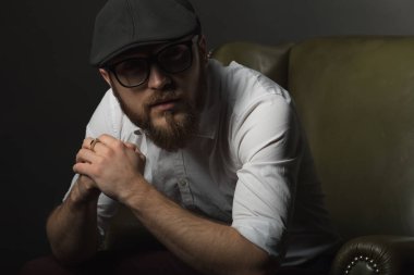 siyah bir kap içinde sakal ve bıyık giyen genç yakışıklı bir adam ve beyaz bir gömlek bir deri yeşil koltuk hamile oturan ve karanlık bir stüdyoda elleriyle jestleri gösteren