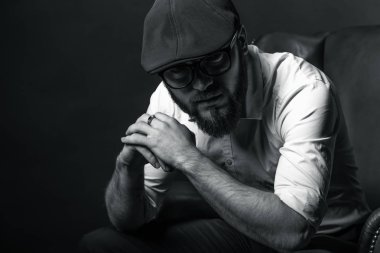 siyah bir kap içinde sakal ve bıyık giyen genç yakışıklı bir adam ve beyaz bir gömlek bir deri yeşil koltuk hamile oturan ve karanlık bir stüdyoda elleriyle jestleri gösteren