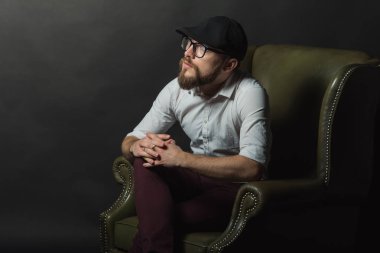 siyah bir kap içinde sakal ve bıyık giyen genç yakışıklı bir adam ve beyaz bir gömlek bir deri yeşil koltuk hamile oturan ve karanlık bir stüdyoda elleriyle jestleri gösteren