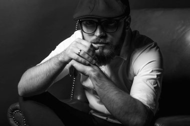 siyah bir kap içinde sakal ve bıyık giyen genç yakışıklı bir adam ve beyaz bir gömlek bir deri yeşil koltuk hamile oturan ve karanlık bir stüdyoda elleriyle jestleri gösteren