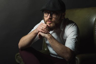 siyah bir kap içinde sakal ve bıyık giyen genç yakışıklı bir adam ve beyaz bir gömlek bir deri yeşil koltuk hamile oturan ve karanlık bir stüdyoda elleriyle jestleri gösteren