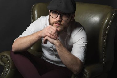 siyah bir kap içinde sakal ve bıyık giyen genç yakışıklı bir adam ve beyaz bir gömlek bir deri yeşil koltuk hamile oturan ve karanlık bir stüdyoda elleriyle jestleri gösteren