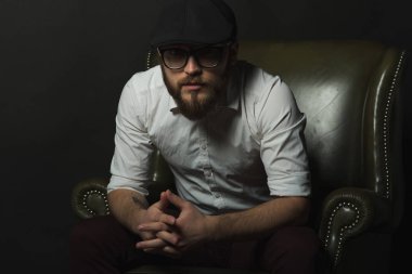 siyah bir kap içinde sakal ve bıyık giyen genç yakışıklı bir adam ve beyaz bir gömlek bir deri yeşil koltuk hamile oturan ve karanlık bir stüdyoda elleriyle jestleri gösteren