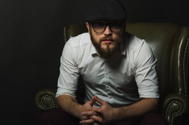 siyah bir kap içinde sakal ve bıyık giyen genç yakışıklı bir adam ve beyaz bir gömlek bir deri yeşil koltuk hamile oturan ve karanlık bir stüdyoda elleriyle jestleri gösteren