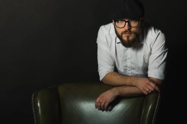 siyah bir kap içinde sakal ve bıyık giyen genç yakışıklı bir adam ve beyaz bir gömlek, koyu bir stüdyoda bir deri yeşil sandalyenin arkasına eğildi