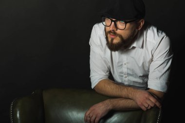 siyah bir kap içinde sakal ve bıyık giyen genç yakışıklı bir adam ve beyaz bir gömlek, koyu bir stüdyoda bir deri yeşil sandalyenin arkasına eğildi