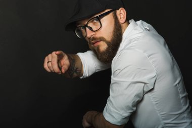 siyah bir kap içinde sakal ve bıyık giyen genç yakışıklı bir adam ve beyaz bir gömlek, koyu bir stüdyoda bir deri yeşil sandalyenin arkasına eğildi