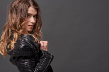 Genç, narin, seksi, Avrupalı, dalgalı saçlı, deri ceketli ve kot pantolonlu bir kız. Gri bir arkaplanda