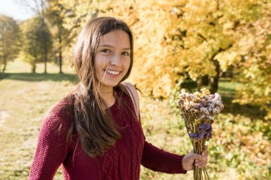 Genç, sempatik Avrupalı görünümlü, kiraz rengi süveter giyen bir kız gülümsüyor, arka planda antik bir kale, kolonlar, sonbahar ılık sabahı. 
