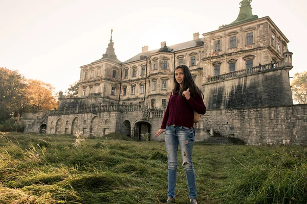 The young pretty sympathetic European appearance of a girl in a cherry sweater and jeans bounces on the back of the old castle. Autumn warm sunny morning