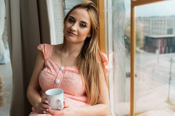 若い金髪の女の子が窓のそばに座ってコーヒーを飲みながら外で雨が降って春の天気 — ストック写真