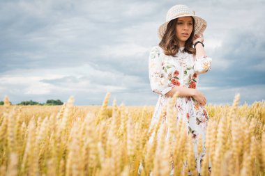 Sarışın, uzun saçlı, yüzünde mutlu bir gülümsemeyle genç bir kızın portresi, düz görünüyor, şapkasını düzeltiyor, açık renk bir elbise ve açık renk bir şapka giyiyor, yaz ya da sonbahar otlaklarında ayakta duruyor.