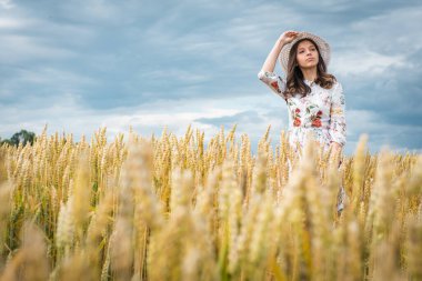 Sarışın, uzun saçlı, yüzünde mutlu bir gülümsemeyle genç bir kızın portresi, düz görünüyor, şapkasını düzeltiyor, açık renk bir elbise ve açık renk bir şapka giyiyor, yaz ya da sonbahar otlaklarında ayakta duruyor.