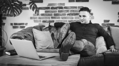 Çoklu görev. Siyah gömlekli ve kotlu yakışıklı bir adam kanepede uzanıyor, yanında masanın üstünde dizüstü bilgisayarı açık, ofiste çalışıyor. Uzaktan kumanda. Siyah beyaz fotoğraf