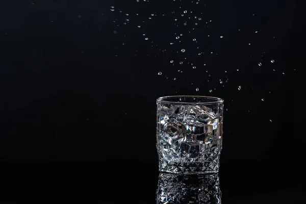 Low key. An ice cube falls into a glass of clear water, on a dark background. Liquid cooling concept.