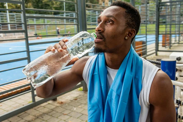 Young muscular build man drinking water bottle after running, attractive athlete resting after outdoor workout, fitness and healthy lifestyle concept.