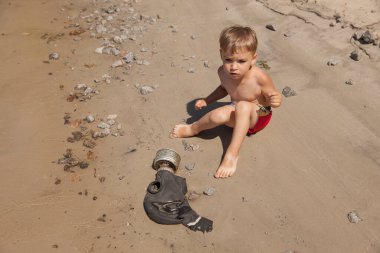 Küçük çocuk nehir kıyısında eski bir gaz maskesinin yanında oturuyor.