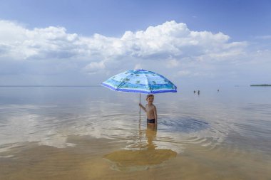 Küçük bir çocuk bir plaj şemsiyesinin altında suda duruyor.