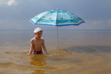 Küçük bir çocuk bir plaj şemsiyesinin altında suda duruyor.