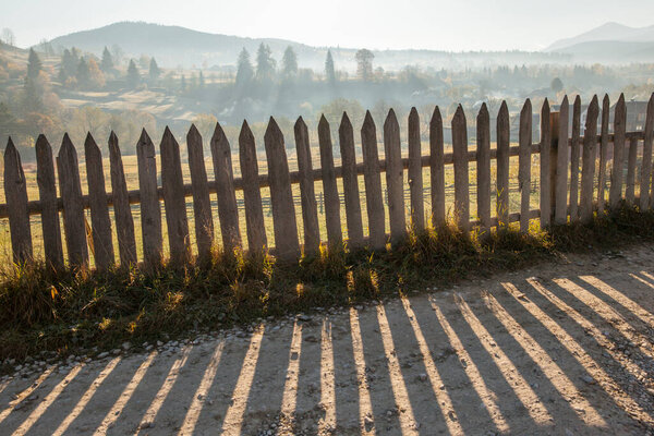 A wooden fence and a shadow from it on a dirt road against the background of the rising sun over the mountains. Carpathians. Ukraine