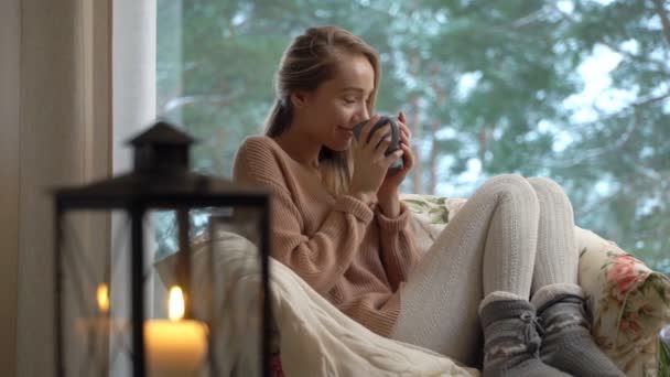 Joven mujer feliz disfrutar de la taza de café caliente sentado en casa por la ventana grande con fondo de árbol de nieve de invierno — Vídeo de stock