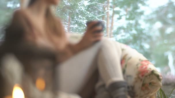 Молода щаслива жінка насолоджується чашкою гарячої кави, що сидить біля великого вікна з зимовим фоном снігового дерева — стокове відео