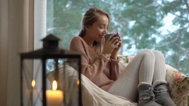 若い幸せな女は冬雪ツリーの背景色の付いた大きなウィンドウでホームに座ってホット コーヒーのカップを楽しむ — ストック動画