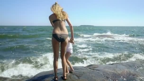 Une jeune femme et sa fille marchant et jouant avec les vagues de la mer parmi les rochers côtiers — Video