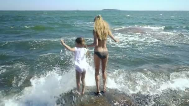 Genç bir kadın ve kızı yürüyüş ve kıyı kayaların arasındaki deniz dalgaları ile oynarken — Stok video