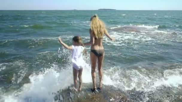 Uma jovem mulher e sua filha andando e brincando com as ondas do mar entre as rochas costeiras — Vídeo de Stock