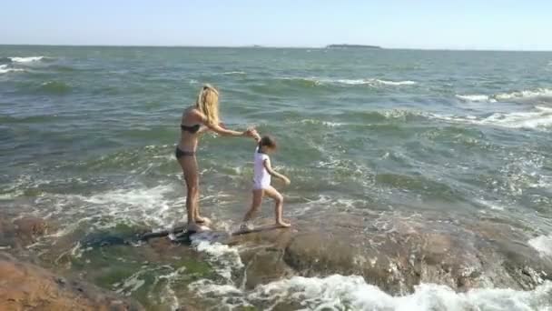 Eine junge Frau und ihre Tochter spazieren und spielen mit den Meereswellen zwischen den Küstenfelsen — Stockvideo
