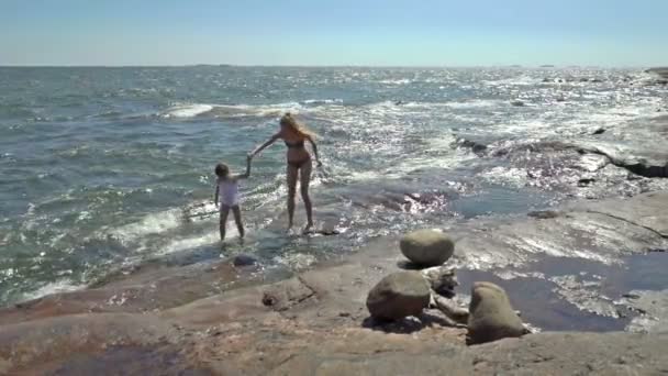 Una joven y su hija caminando y jugando con las olas del mar entre las rocas costeras — Vídeo de stock