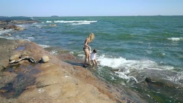 Uma jovem mulher e sua filha andando e brincando com as ondas do mar entre as rochas costeiras — Vídeo de Stock