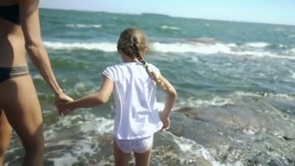 Een jonge vrouw en haar dochter wandelen en spelen met de golven van de zee tussen de kust rotsen — Stockvideo